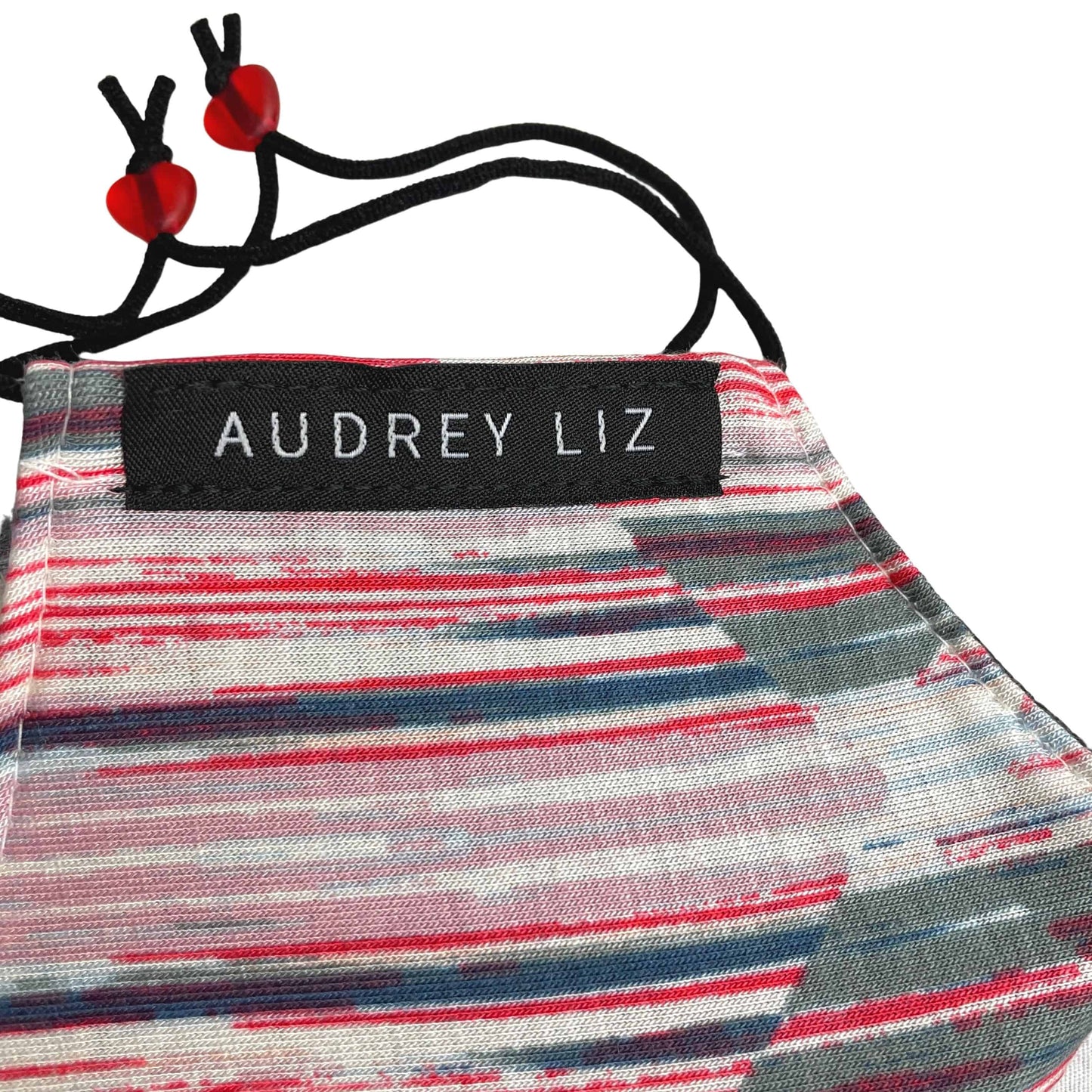 Detailed view of women’s cloth face mask in a multi-color stripe pattern, with rectangle Audrey Liz logo sewn on side.