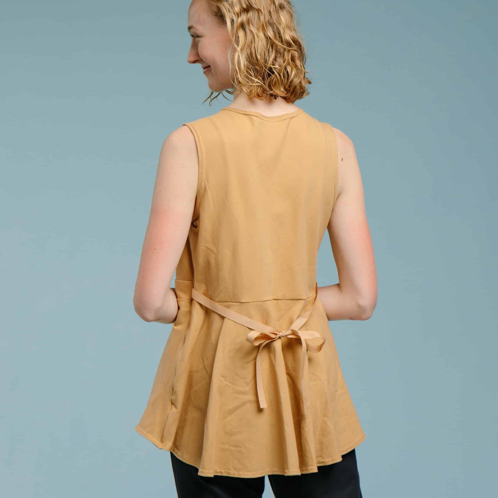 Back view of model wearing a tan brown mastectomy shirt that has an adjustable ribbon that ties in the back to accommodate various bust sizes.