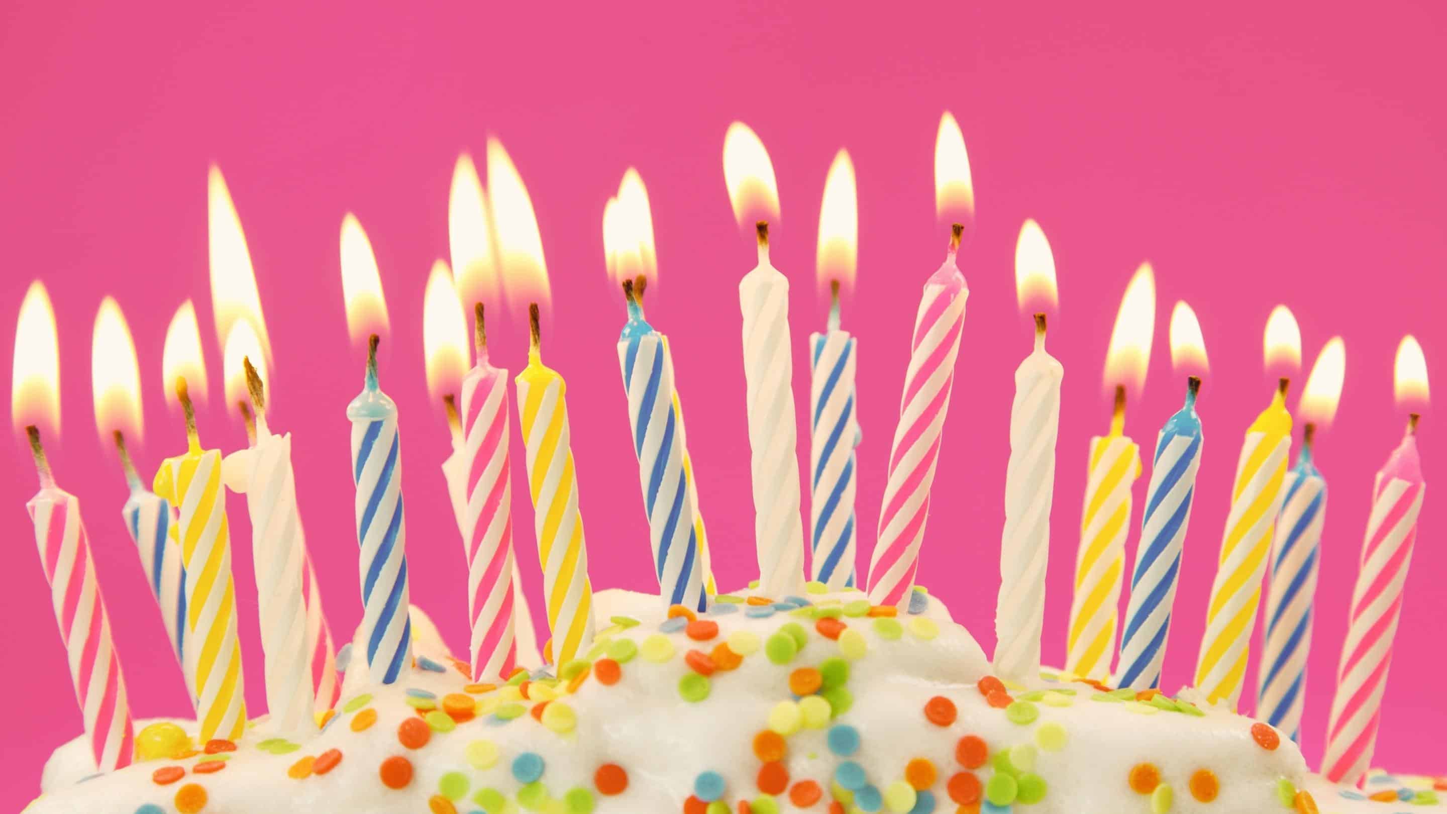 Colorful birthday cake with numerous candles lit on a pink background.
