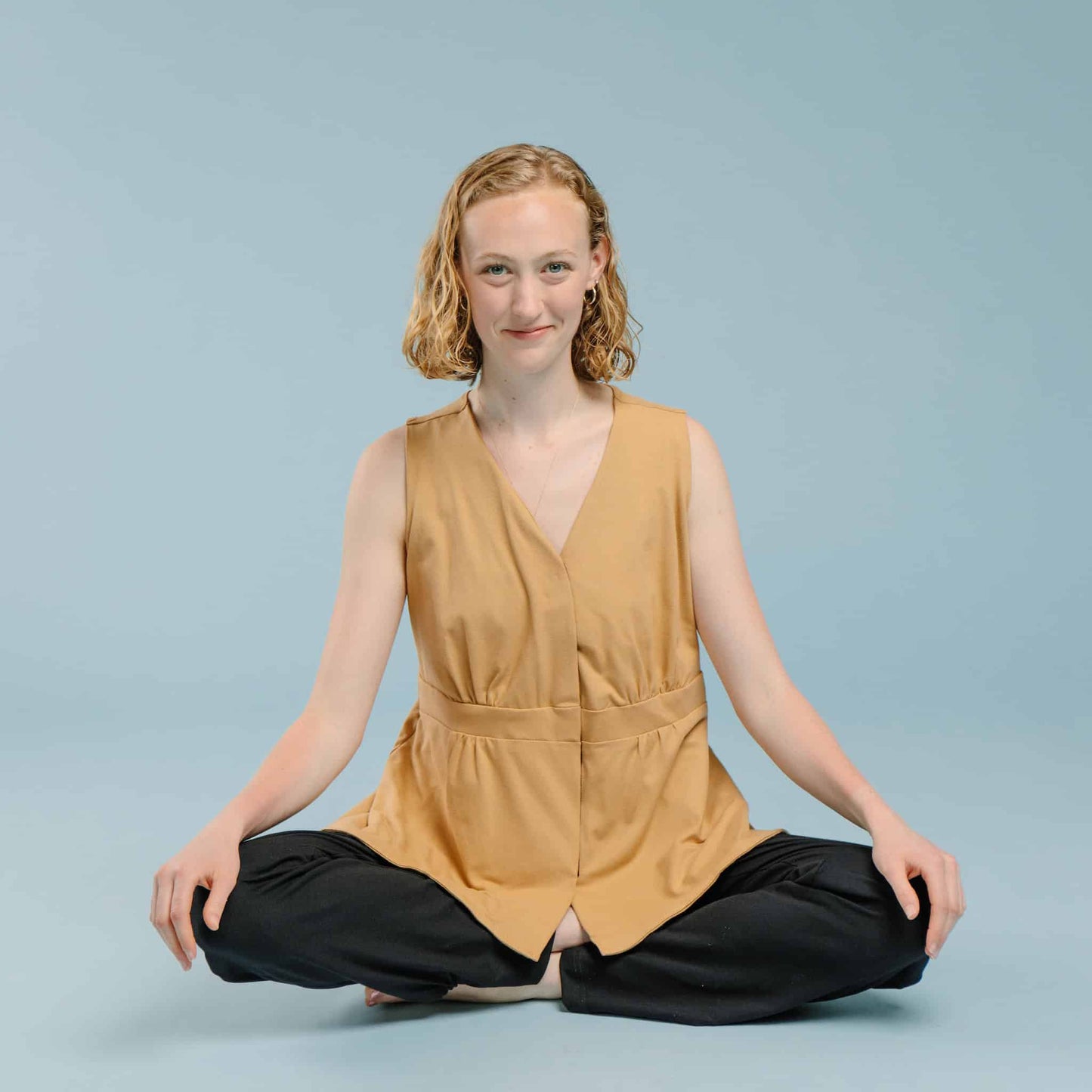 Woman wearing a tan sleeveless mastectomy shirt, with velcro front closure and flattering pleats above and below waistband.