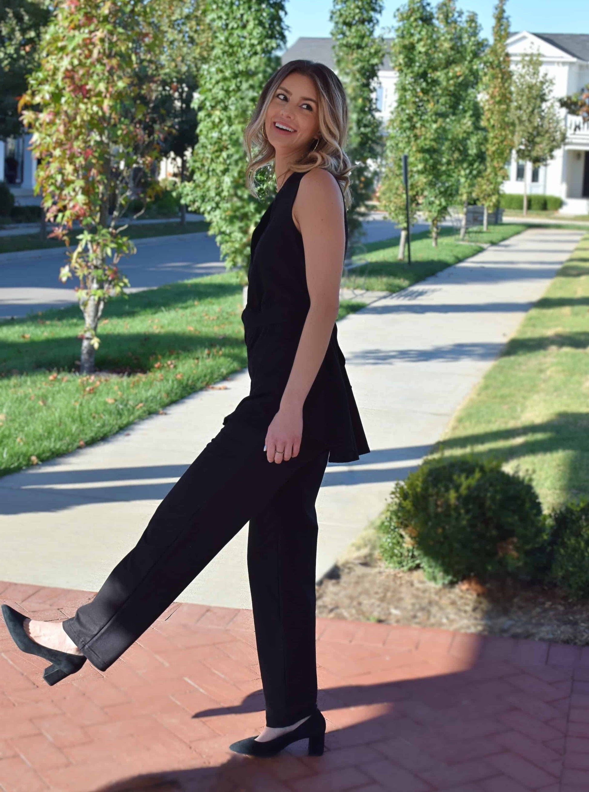 Smiling woman wearing a black Audrey Liz post-mastectomy shirt and coordinating pants. Styled with heels standing on a sunny sidewalk.