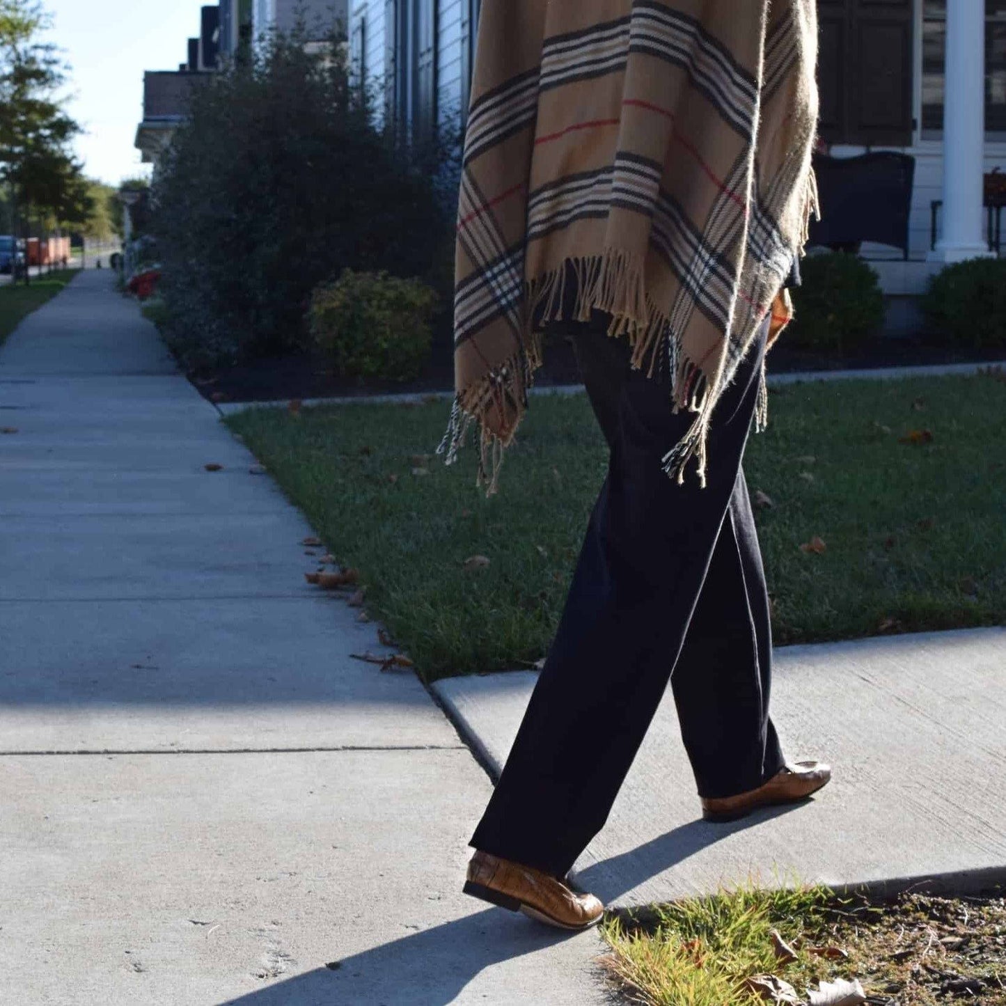 Person walking on a sidewalk wearing black pull-on pants, styled with brown loafers and plaid poncho.