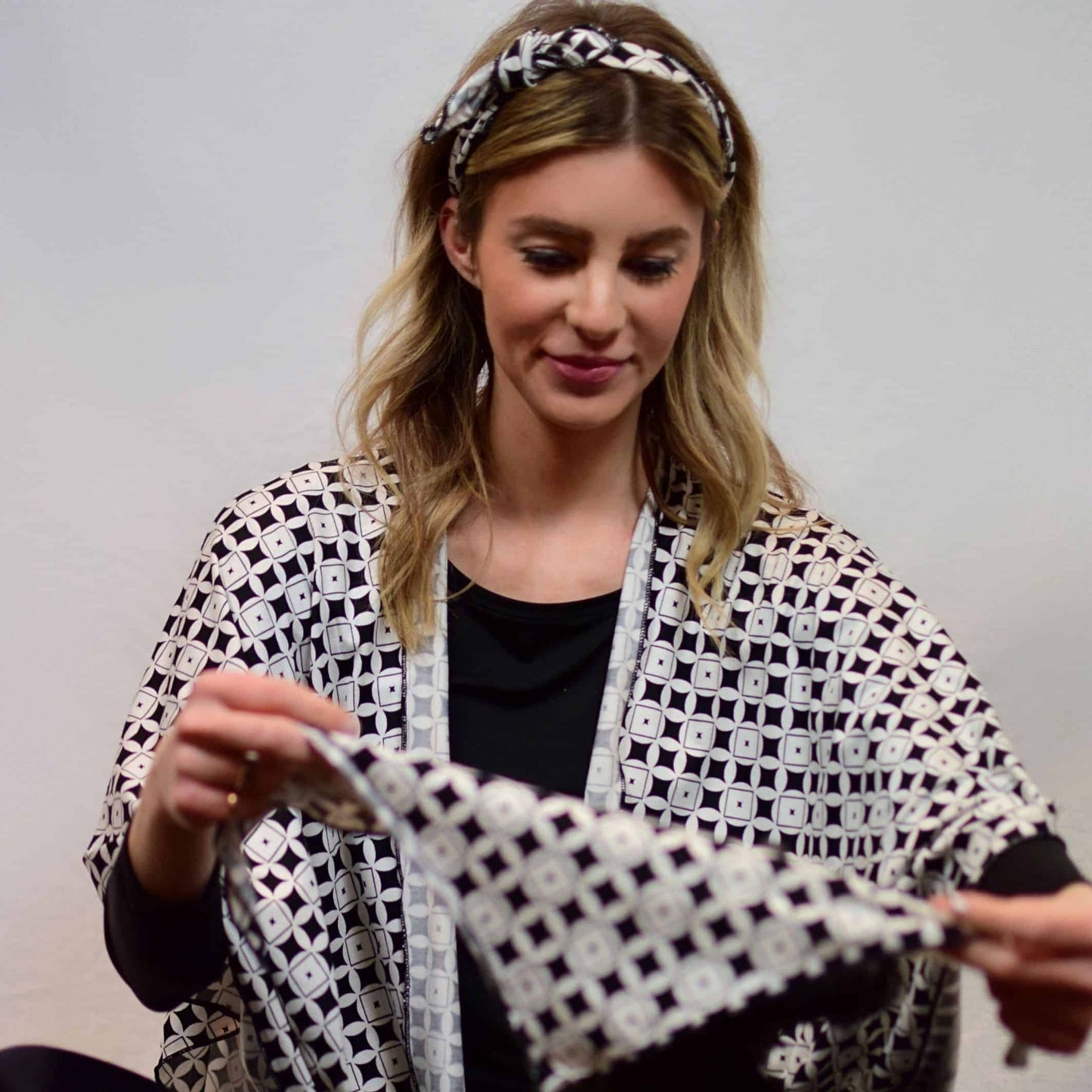 Woman sitting, wearing a black and white print shawl with a matching bandana worn as a headband, folding a smaller bandana with hands to place on dog.