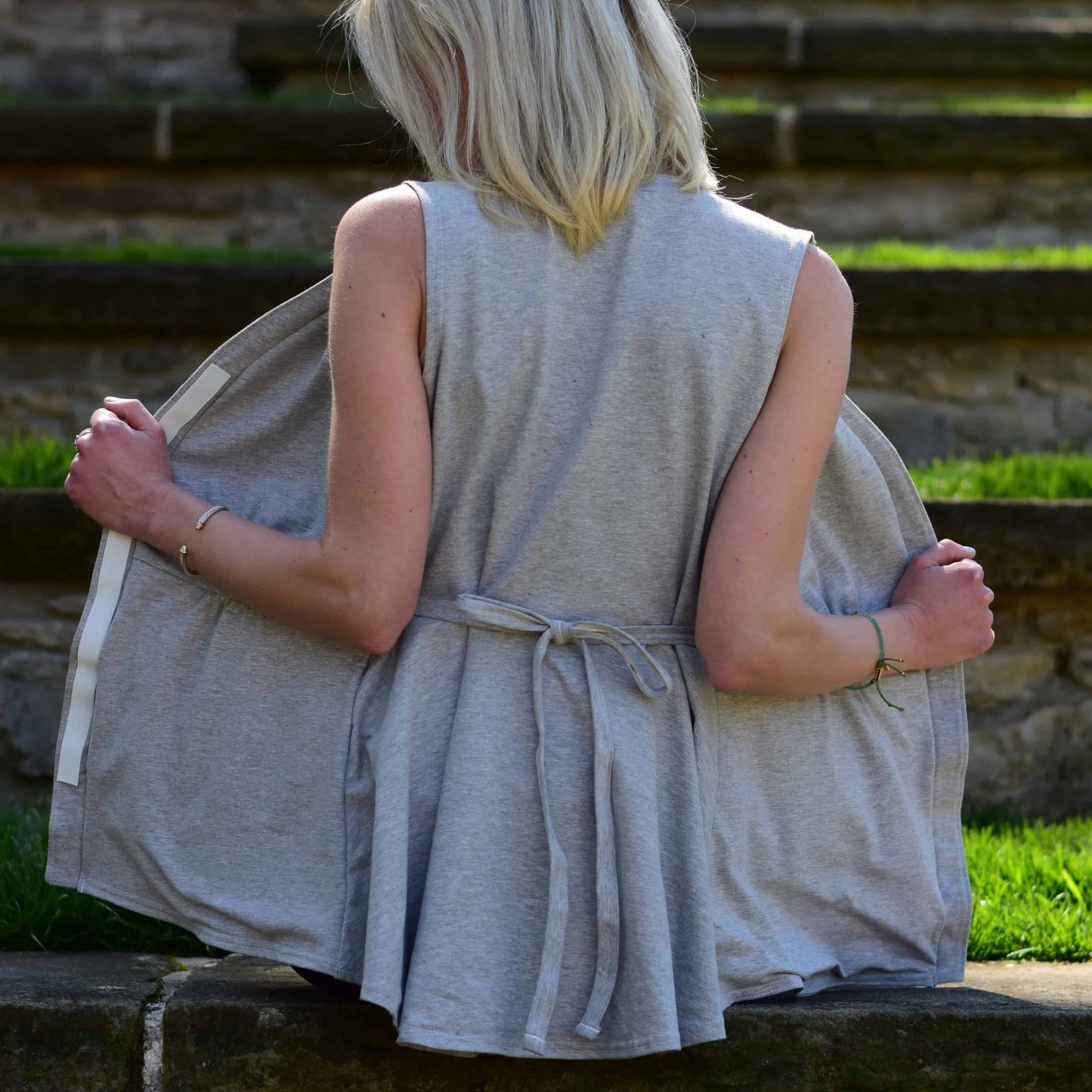 Woman sitting in a shaded outdoor area, holding her top open, with only the back details of the shirt visible.