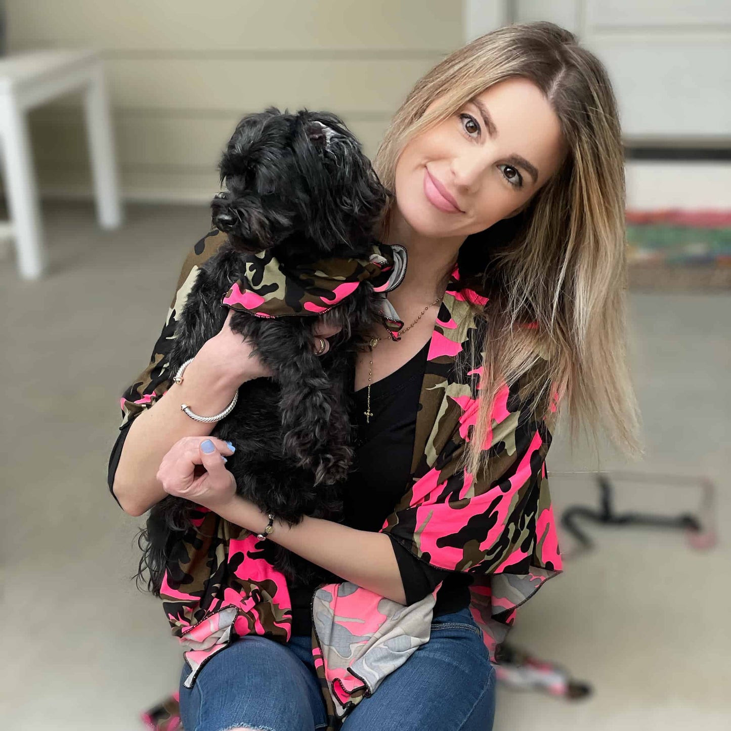 Woman holding a small black dog sitting on concrete steps. She wears a pink camo print shawl over shoulders, while dog wears a bandana around neck in matching camo pattern.