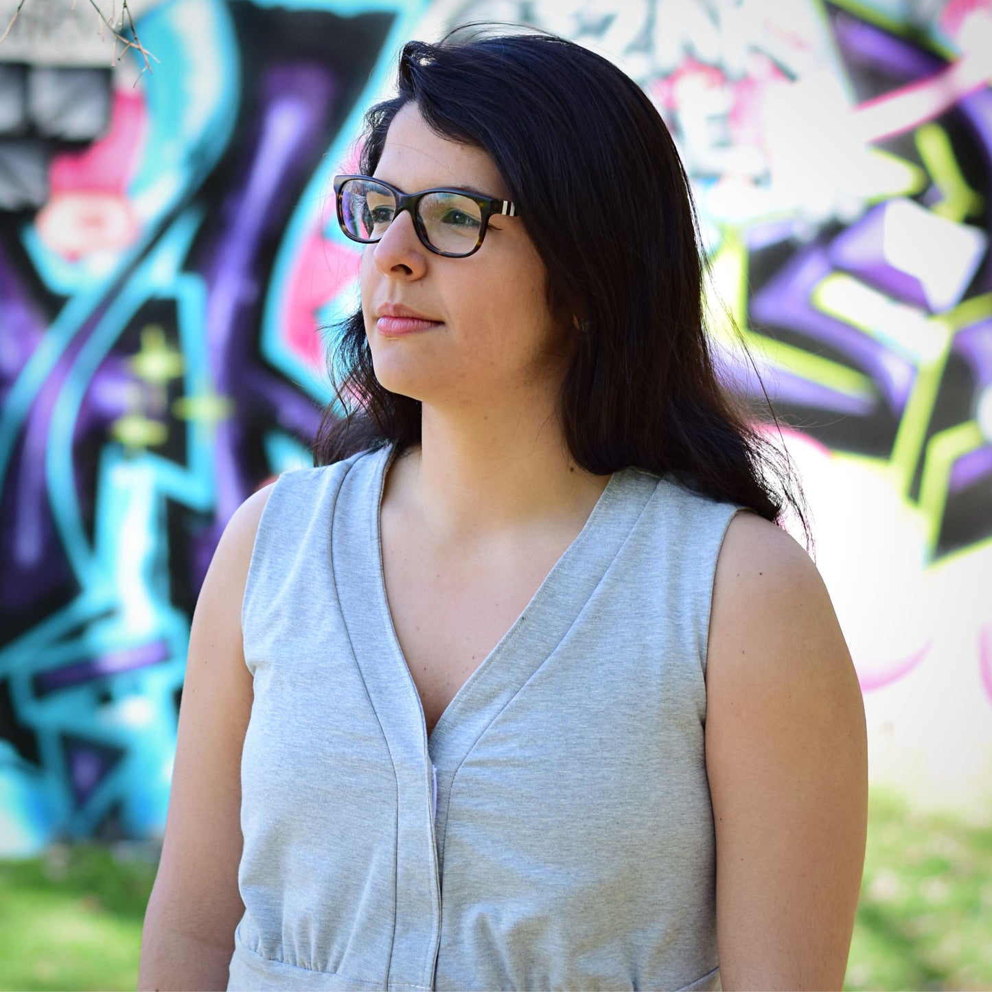 Woman wearing a heather grey v-neckline top, standing outdoors with a blurred painted wall in the background.